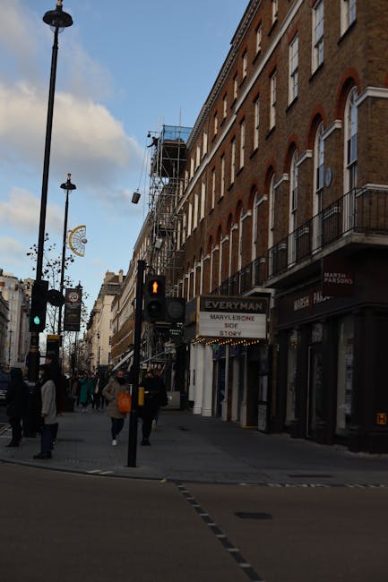 A busy street scene in Marylebone showing a multi-storey brick building with large windows and small balconies on the right side, and scaffolding on its side indicating building work. Pedestrians are walking along the pavement, some carrying shopping bags. Traffic lights are visible at the intersection, with a red light illuminated. Several street lamps line the street, and shop signs, including one for 'EVERYMAN,' are displayed on the ground level of the building. The scene suggests an urban environment suitable for house removals and furniture transport services, with loose cobblestones and a clear sky overhead.