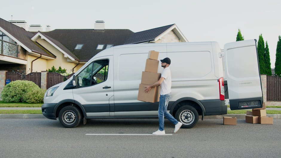 A man dressed in casual clothing and wearing a face mask is loading three cardboard boxes into the side of a white delivery van parked on a residential street. The van is positioned parallel to the curb in front of a property with a dark sloped roof, skylights, and a cream-colored exterior. The man is carrying two of the boxes, one under each arm, while a third box is stacked on top of the others. Several additional cardboard boxes are placed on the pavement nearby, indicating a home relocation or furniture transport process managed by Man with Van Marylebone. The environment includes well-maintained greenery, such as bushes and tall trees, and the scene is lit with natural daylight, showing a clear and calm day suitable for packing and moving activities.