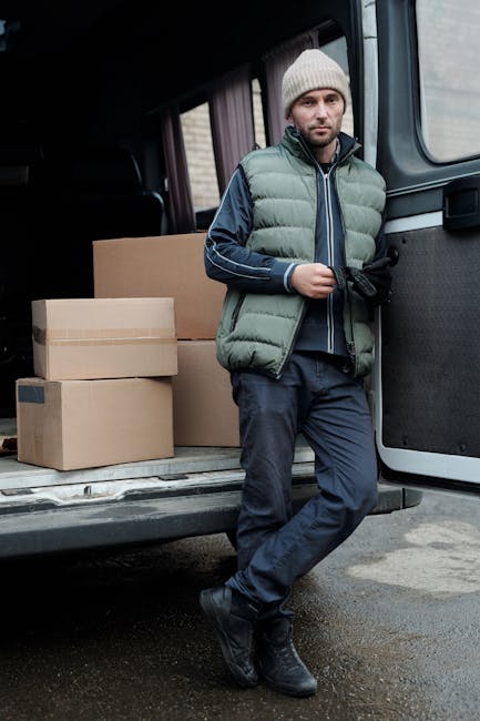 A man wearing a grey knitted beanie, a green padded vest over a dark blue hoodie, and dark trousers stands next to an open van door during a home relocation process. Inside the van, several cardboard boxes of varying sizes are visible, some stacked. The man is holding a black glove or tool in his hand and appears to be preparing for or taking a break from loading or unloading items. The interior of the van shows a dark space, with the open rear door revealing a pavement outside. This scene illustrates furniture transport and packing activities involved in professional removals, with the company Man with Van Marylebone providing move and relocation services, supporting efficient home moving logistics.