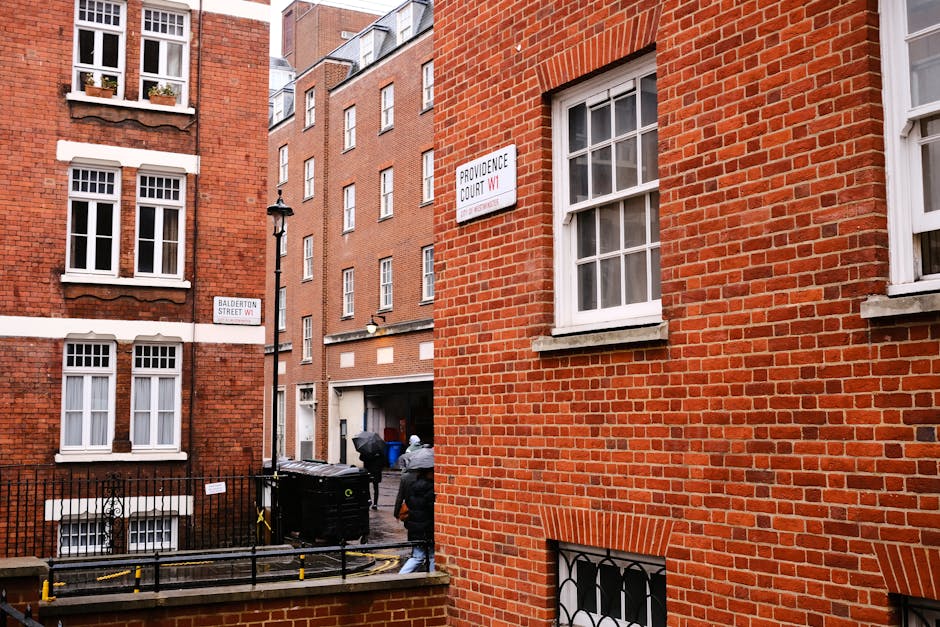 The image shows the exterior view of a red brick building situated at the corner of Balderton Street and Providence Court in Marylebone, London. The building features multiple white-framed sash windows on each floor, with some windows displaying flower boxes. A black metal fence lines the sidewalk below the windows, and a black trash bin is positioned against the fence. Two individuals, one wearing a grey jacket and the other in dark clothing, are seen carrying large cardboard boxes or packing materials out of the building, suggesting active packing or home relocation preparations. The scene takes place during daylight with natural lighting illuminating the brick facades and street signs. This setting reflects a typical urban environment where furniture transport and packing are part of a residential moving process, an area serviced by companies like Man with Van Marylebone as they assist with removals and moving logistics.