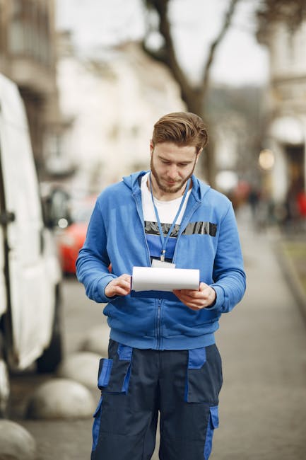 A man wearing a blue branded uniform with reflective strips on the chest and pockets stands outdoors on a city street during daytime, holding a clipboard and reviewing documents. Behind him, blurred vehicles are parked along the roadside, and trees with some foliage are visible, indicating a typical urban environment. This scene suggests he is involved in planning or overseeing a home relocation or removal project managed by Man with Van Marylebone, which specialises in house removals and moving services. The lighting is natural, and the man appears focused and attentive, possibly coordinating the loading process or ensuring correct procedures are followed in the context of furniture transport or packing and moving logistics.