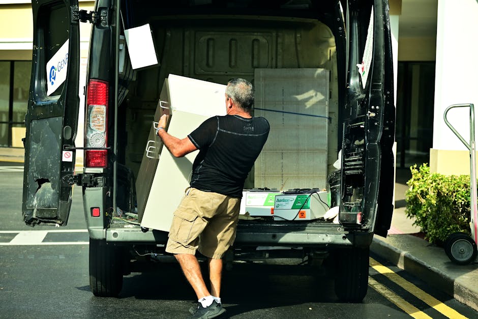 A man with gray hair, wearing a black T-shirt, beige cargo shorts, and black sneakers, is loading a large white cabinet into the back of a black moving van parked on a city street. The van's rear doors are fully open, revealing protective blankets and packing materials inside to secure the furniture during transport. A green box of disposable gloves is also visible inside the van. The man is positioned on the pavement, lifting the cabinet with both hands, with his back to the camera. To the right, part of a green shrub and a hand truck are visible near the curb. The background includes a modern glass building with reflections of the street scene. This scene depicts a typical home relocation process involving furniture transport, with the team from Man with Van Marylebone managing packing and loading for a smooth house removal.