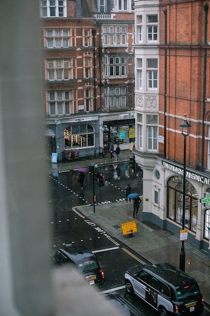 View of a busy street scene on Marylebone High Street seen from an elevated position, with wet pavement indicating recent rain. In the foreground, there is a white building with a curved corner and a visible sign for a medical practice, along with a bicycle rack and a street lamp. Across the street, an alleyway serves as a loading bay area where boxes, furniture wrapped in plastic, and moving blankets are being carefully loaded or unloaded by movers from Man with Van Marylebone. A van is parked nearby, demonstrating the final stages of a home relocation or furniture transport process. Pedestrians with umbrellas are walking along the street, and the surrounding multi-storey buildings with traditional brick facades and sash windows frame the scene. The environment suggests an urban setting suited for professional removals and moving services, with clear signs of packing and loading activities taking place in and around the loading bay area.