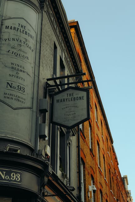 Photograph of the exterior of a historic building on Marylebone High Street, featuring two hanging shop signs labeled 'The Marylebone' along with their address details. The building has a curved corner facade with decorative molding, and its weathered brickwork is visible alongside a large window. The signs are attached to black metal brackets extending from the building's facade. The sky above is clear and blue, indicating daytime. While this image does not show interior scenes, it contextualizes the setting where home relocation and furniture transport services by Man with Van Marylebone may take place, especially in relation to loading bay rules and access points for removals or moving logistics.