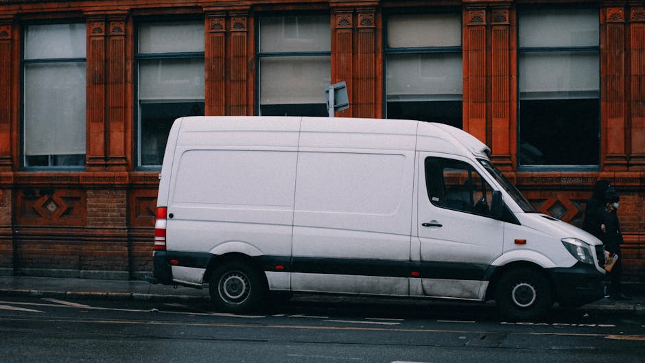 A white commercial van parked parallel to a red brick building with decorative architectural details, situated on a city street with double yellow lines. The van's side door is closed, and the vehicle appears to be in the process of being loaded or unloaded as part of a house removal or furniture transport operation. Behind the van, a person dressed in dark clothing is walking along the pavement. The building features large windows with white frames and intricate brickwork patterns above and below the windows. The scene is lit with natural daylight, emphasizing the urban environment and the vehicle's role in relocation services, such as packing and moving for property owners in Marylebone managed by Man with Van Marylebone.