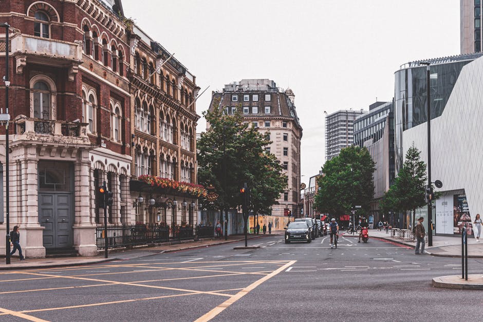 A view of a busy street in Marylebone, London, showing historic and modern buildings on either side of the road. The left side features a red-brick building with white accents, arched windows, and decorative balconies, with hanging flower baskets and black wrought iron fencing. The right side displays contemporary glass-fronted structures with sleek, curved facades. Several pedestrians walk along the pavement, while cars and cyclists are seen navigating the intersection. A few trees line the street, providing greenery amidst the urban environment. The traffic lights are red, and the sky overhead is overcast, casting diffuse lighting over the scene. This setting illustrates the typical urban environment where home relocation and furniture transport services by Man with Van Marylebone might operate, involving the loading and unloading process of household items with professional removals equipment visible nearby.