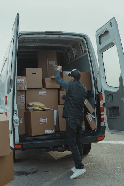 A man dressed in a blue long-sleeve shirt, black padded vest, black trousers, white sneakers, and a grey cap stands beside a white moving van with a brown panel at the back, parked outside a residenti