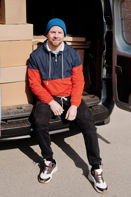 A man wearing a blue beanie hat, a navy and red windbreaker jacket, black pants, and white sneakers is sitting on the open tailgate of a black moving van. Inside the van, there are several cardboard boxes of various sizes, some wrapped in plastic or packing paper, stacked neatly against the interior walls. The boxes are arranged on the ground and appear ready for loading or unloading. The man is outside on a paved surface, smiling at the camera, with natural light illuminating the scene. The van door is fully open, revealing the interior filled with packing materials, and the side of the vehicle is visible on the right. This scene captures a moment during a home relocation process, focusing on the loading or packing phase, with a subtle emphasis on the logistics of furniture transport and packing for a house move, associated with professional removals services by Man with Van Marylebone.