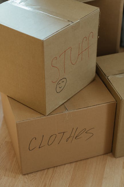 Several cardboard boxes stacked inside a residential property, with one labeled 'STUFF' and another labeled 'CLOTHES' written in black marker. The 'STUFF' box has a simple smiley face drawing beneath the label. The boxes are placed on a wooden floor, indicating a home relocation or packing process. The environment appears to be a room or corridor with natural lighting. These boxes are part of the packing phase typical of house removals, carried out by specialists such as Man with Van Marylebone, who assist with packing, loading, and transportation services to facilitate a smooth moving process.