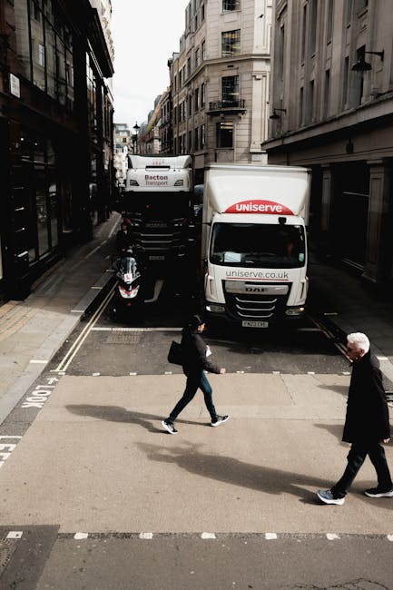 A narrow street in an urban area with tall, multi-storey buildings on both sides. In the foreground, two pedestrians are crossing the street—one woman carrying a backpack and a black bag, and an older man wearing a dark coat. Behind them, a small black scooter is parked on the pavement. Further back, there are two large white moving trucks parked side by side on the street; the trucks display company branding related to home relocation services. The street appears to be in the midst of a moving or delivery process, with the trucks likely used for furniture transport or packing and moving tasks. The scene is well-lit with natural daylight, typical of a city environment, and the urban setting suggests a busy area suitable for professional removals services from Man with Van Marylebone.