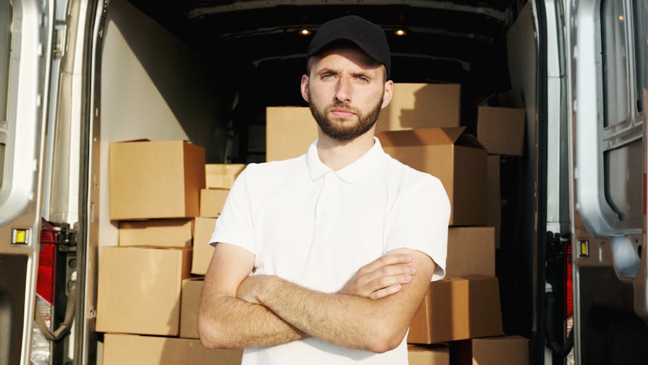 A man wearing a blue beanie and a two-tone jacket with navy blue and bright red sections is sitting on the edge of a black moving van with its side door open, revealing a cargo area filled with neatly stacked cardboard boxes of various sizes. The boxes are secured with plastic wrap and are arranged on the vehicle's floor. The man is smiling and appears to be taking a break during a home relocation process, suggesting a professional moving service such as Man with Van Marylebone. The scene is outdoors on a paved surface, and the lighting indicates daylight conditions. The background shows a partial view of a residential building or urban environment. The image depicts furniture, packaging materials, and the equipment used in packing and loading belongings for a furniture transport or house removal service, supporting the context of professional removals and moving logistics.